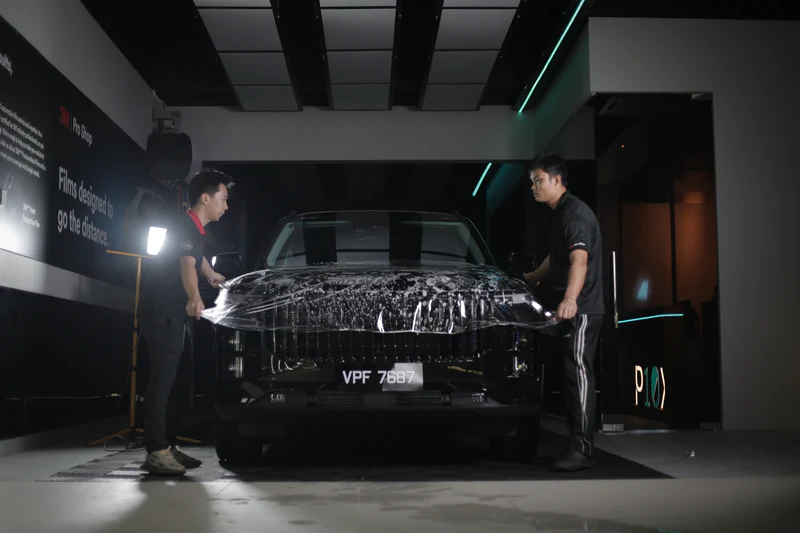 Two men installing a protective film on the hood of a black car inside a dimly lit garage.