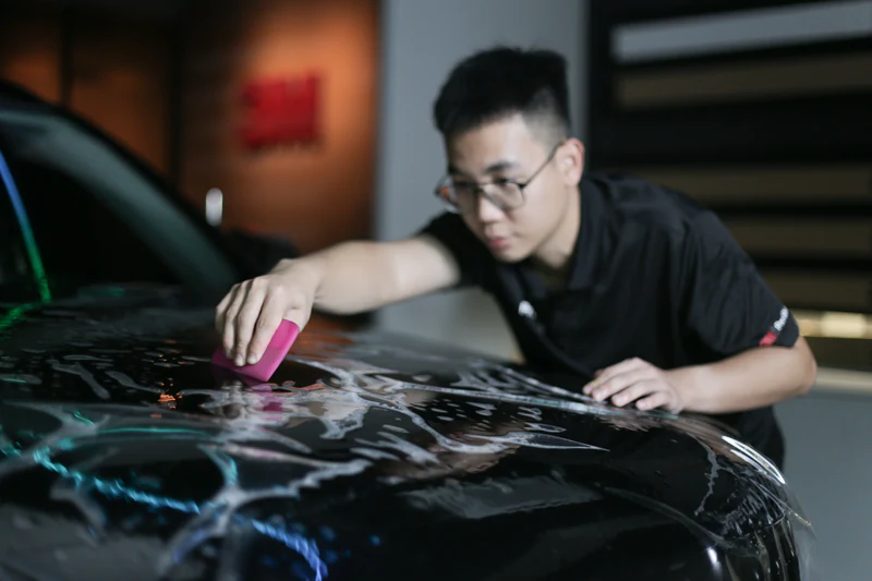 Man wearing glasses carefully cleaning the hood of a black car using a pink sponge with soap suds.