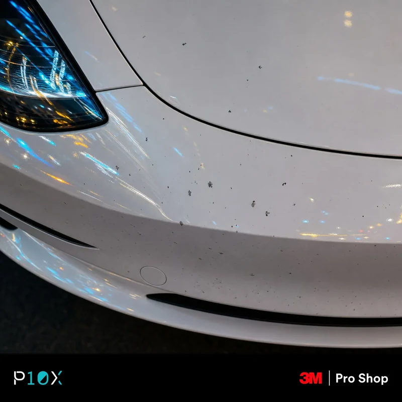 A macro close-up shot of a white Tesla Model 3's front bumper showing small stone chips and paint damage in a Kuala Lumpur urban setting.